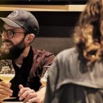 Lachender Mann mit Bart und Brille genießt ein Glas Bier in einer gemütlichen Runde mit Freunden in einer Bar.