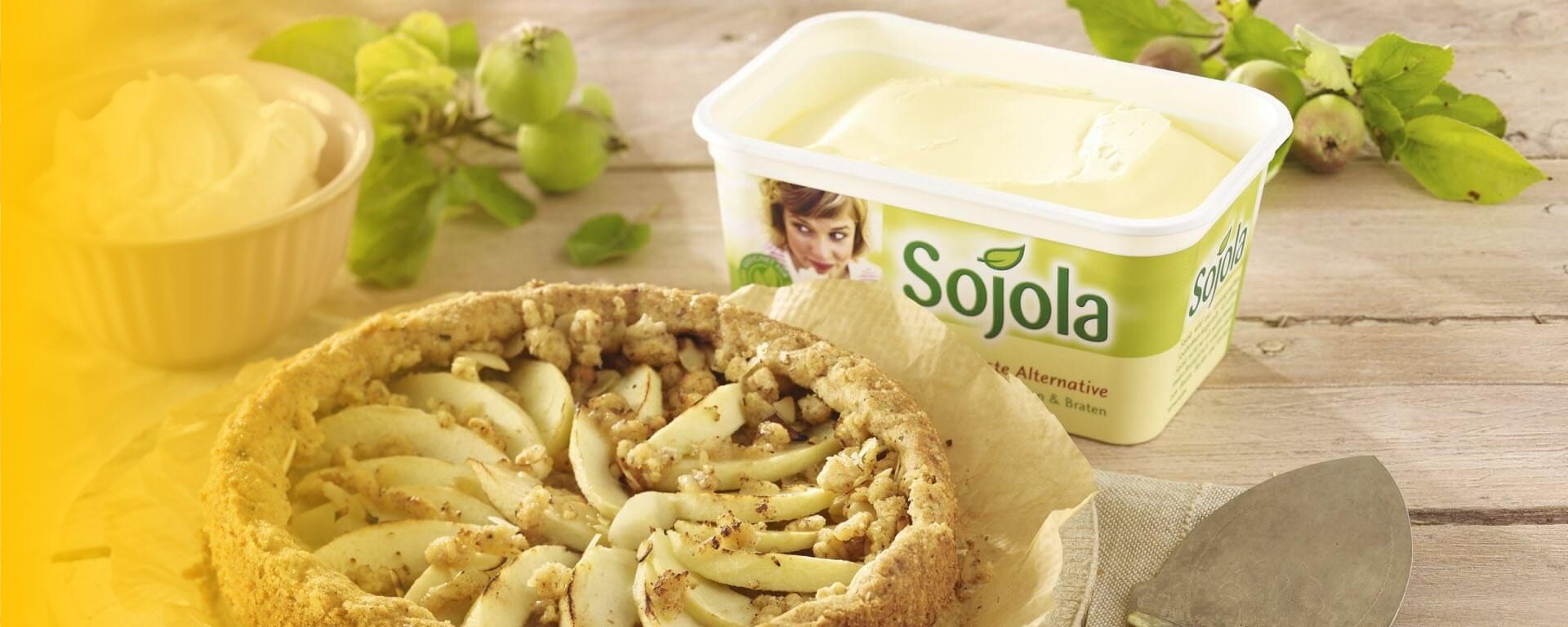 Ein rustikaler Apfelkuchen auf einem Holztisch, neben einer Schale mit Schlagsahne und einer Packung Sojola Margarine, die als pflanzliche Alternative zum Backen verwendet wird. Frische grüne Äpfel und Blätter liegen im Hintergrund und verleihen dem Bild eine natürliche Atmosphäre.