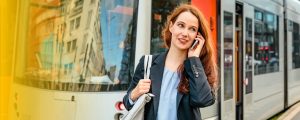 Eine Frau in einem Business-Outfit telefoniert lächelnd mit einem Smartphone und steht vor einer Straßenbahn in der Stadt Düsseldorf. Die Szene zeigt städtische Mobilität und berufliche Vernetzung im Alltag.