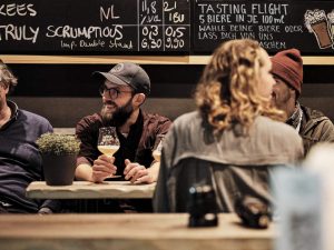 A man in a relaxed bar setting holding a glass of craft beer, surrounded by friends. In the background, a chalkboard displays beer offerings. Symbolic of modern gastronomy and the urban vibe of the New Altstadt.